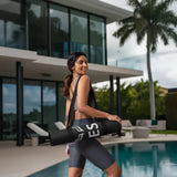 Woman carrying Velite Armor Premium Yoga Mat by a modern poolside, showcasing its sleek black design and portability for active lifestyles.