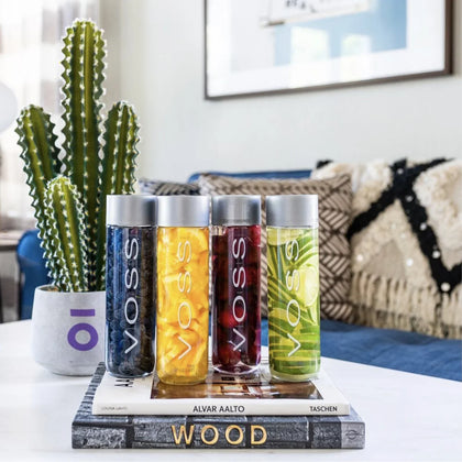 VOSS flavored water bottles infused with fruit and herbs, displayed on books with a cactus and cozy living room in the background.