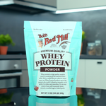 Bob's Red Mill Whey Protein Powder 12 oz pouch displayed in modern kitchen; 14g protein per serving, ideal for shakes and baking, 4-pack option.