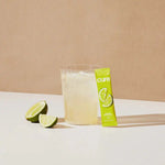 Glass of mixed Cure Hydration Lime Electrolyte drink next to product pouch and lime slices on beige background.