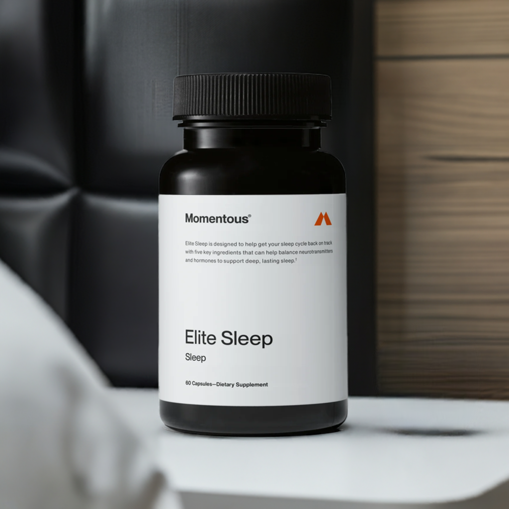Momentous Elite Sleep – suplemento para mejorar la calidad del sueño, con 60 cápsulas diseñadas para equilibrar neurotransmisores y hormonas. Presentado sobre una mesa junto a una cama con fondo en tonos oscuros, resaltando su función para el descanso nocturno.