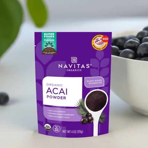 Front view of a purple acai powder pouch placed on a white countertop with greenery in the background.

