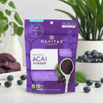 Front view of the acai powder pouch placed on a white table with fruit and plants in the background.

