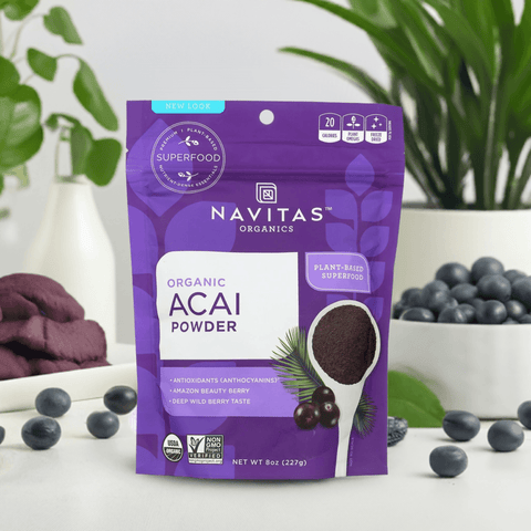 Front view of the acai powder pouch placed on a white table with fruit and plants in the background.

