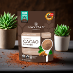 Black cacao powder pouch centered on a counter with plants, cinnamon sticks, and a neutral background.

