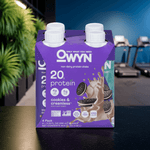 OWYN Cookies & Creamless 20g Plant-Based Protein Shake multipack displayed in a luxury gym setting with tropical plants and workout equipment.