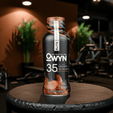 OWYN Pro Elite Protein Shake 35g – Chocolate flavor drink with home gym and indoor cycling bike setup.