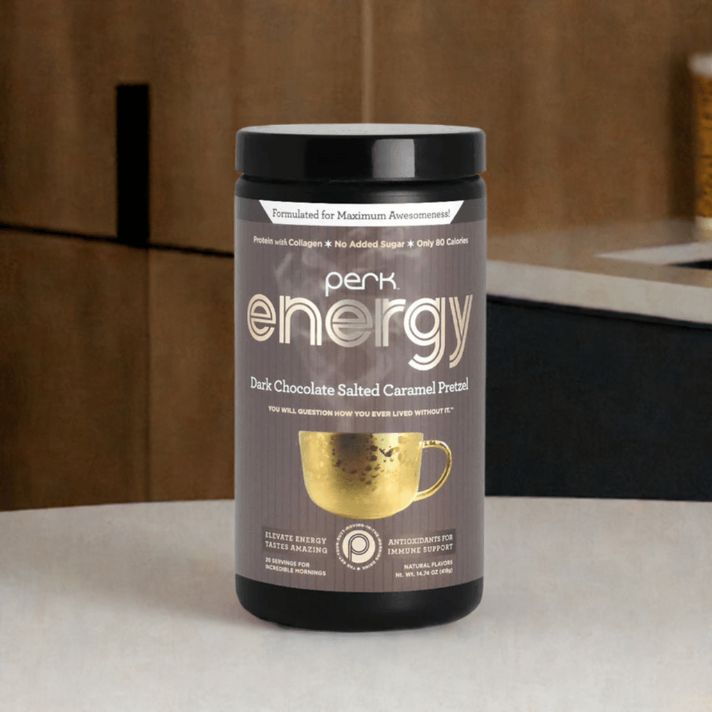 A container of Perk Energy Dark Chocolate Salted Caramel Pretzel sits on a kitchen countertop. The packaging highlights its benefits, including collagen protein, no added sugar, and only 80 calories per serving. The sleek black and gold design emphasizes its premium quality, with a gold mug illustration adding a cozy touch. The background features modern kitchen cabinetry with warm lighting.