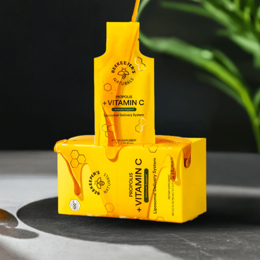 Beekeeper’s Naturals Propolis + Vitamin C – suplemento con sistema de entrega liposomal para el soporte inmunológico. Empaque amarillo vibrante con diseño de miel derramándose, resaltando su ingrediente natural clave. Presentado en un entorno elegante con un fondo de plantas verdes