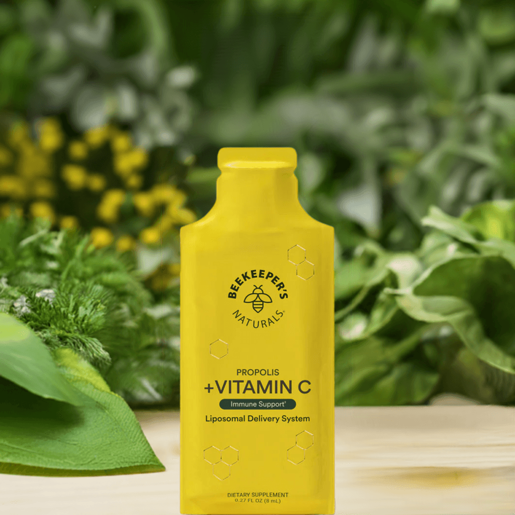 Beekeeper’s Naturals Propolis + Vitamin C en un entorno natural con plantas verdes. Suplemento en presentación líquida con sistema de entrega liposomal para una absorción óptima y beneficios antioxidantes.
