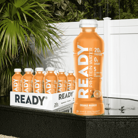 Ready Protein Water Orange Mango bottle on outdoor surface with green plant background

