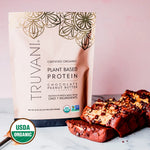 Truvani Organic Chocolate Peanut Butter Protein powder pouch on a table with dates and chocolate chunks, USDA Organic label visible.