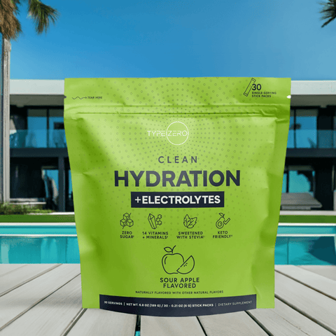 Front view of Clean Hydration + Electrolytes green pouch placed on a wooden deck by a pool, with a modern villa and palm trees in the background.