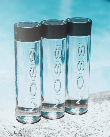Three 850ml glass bottles of VOSS Still Water displayed on ice with a bright blue water backdrop, highlighting purity and elegance.