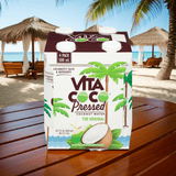 Vita Coco Coconut Water with real coconut pulp in 4-pack, photographed on tropical wooden deck by ocean.