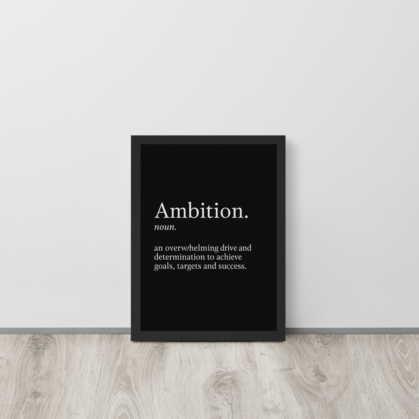 Framed inspirational wall print with the word "Ambition" placed on the floor against a white wall in a minimalist room.