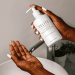 Velite Essentials Lotion - Vegan Hand Body Care held over sink, demonstrating convenient pump dispenser for daily skincare use.