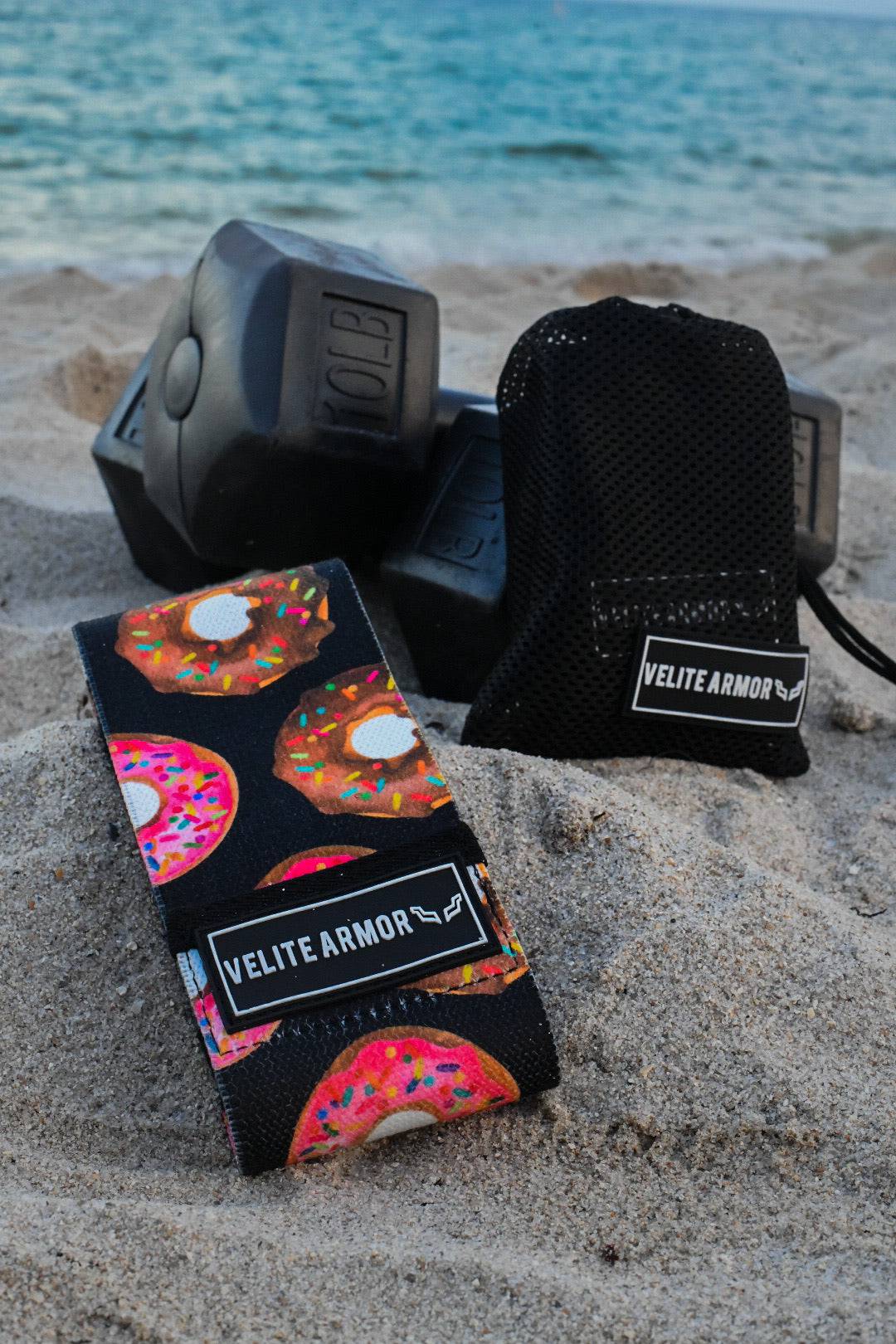 Velite Armor wrist wraps with a vibrant donut pattern, placed on the sand alongside black dumbbells and a mesh carrying pouch, with the ocean in the background. A perfect blend of fun design and serious performance for weightlifting and strength training. 