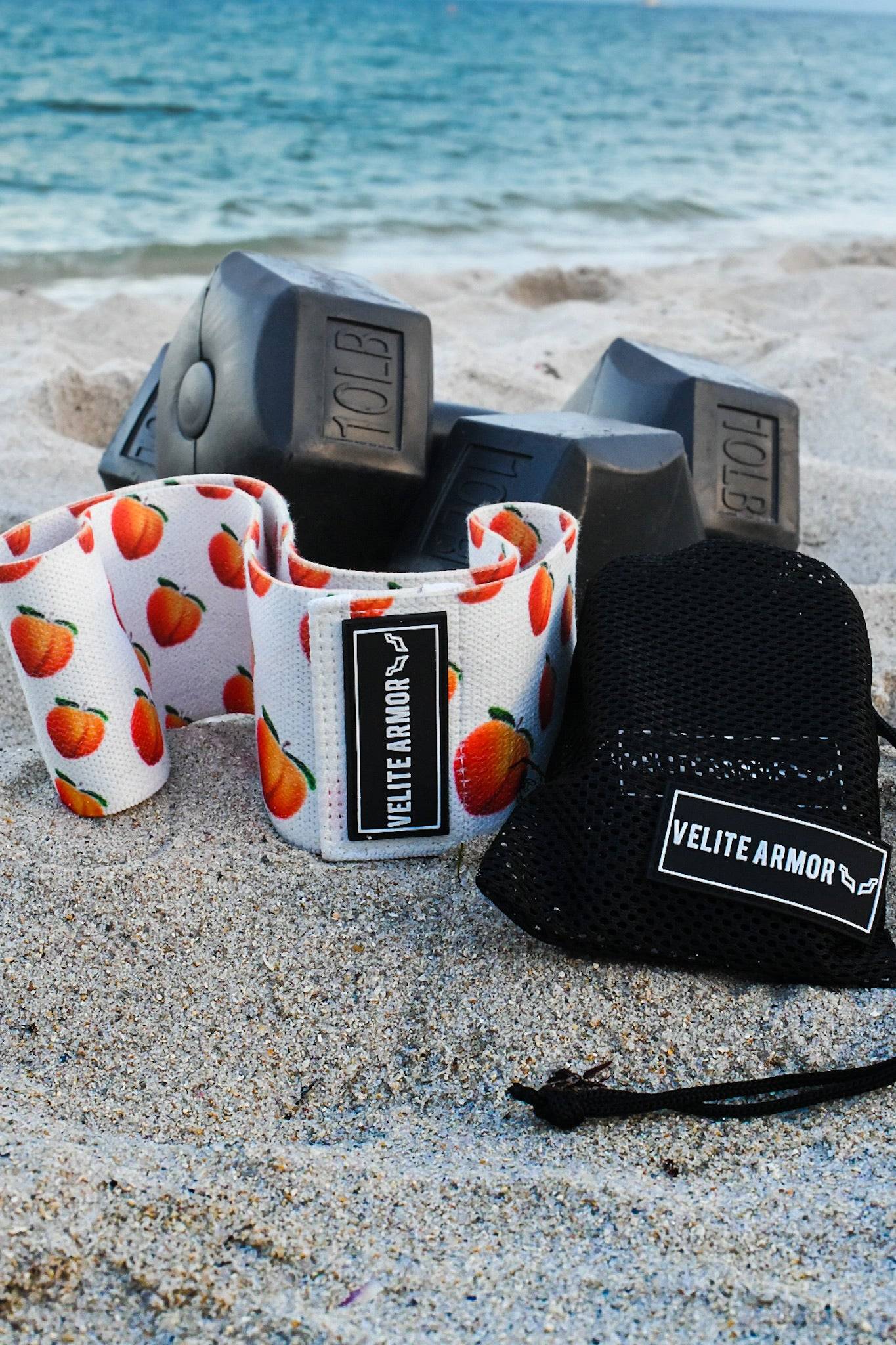 Velite Armor wrist wraps and mesh carrying pouch placed on the sand with black dumbbells in the background, overlooking the ocean. The wrist wraps feature a vibrant peach pattern on white fabric, symbolizing a fusion of style and performance. Perfect for weightlifting and strength training enthusiasts who embrace an active lifestyle.