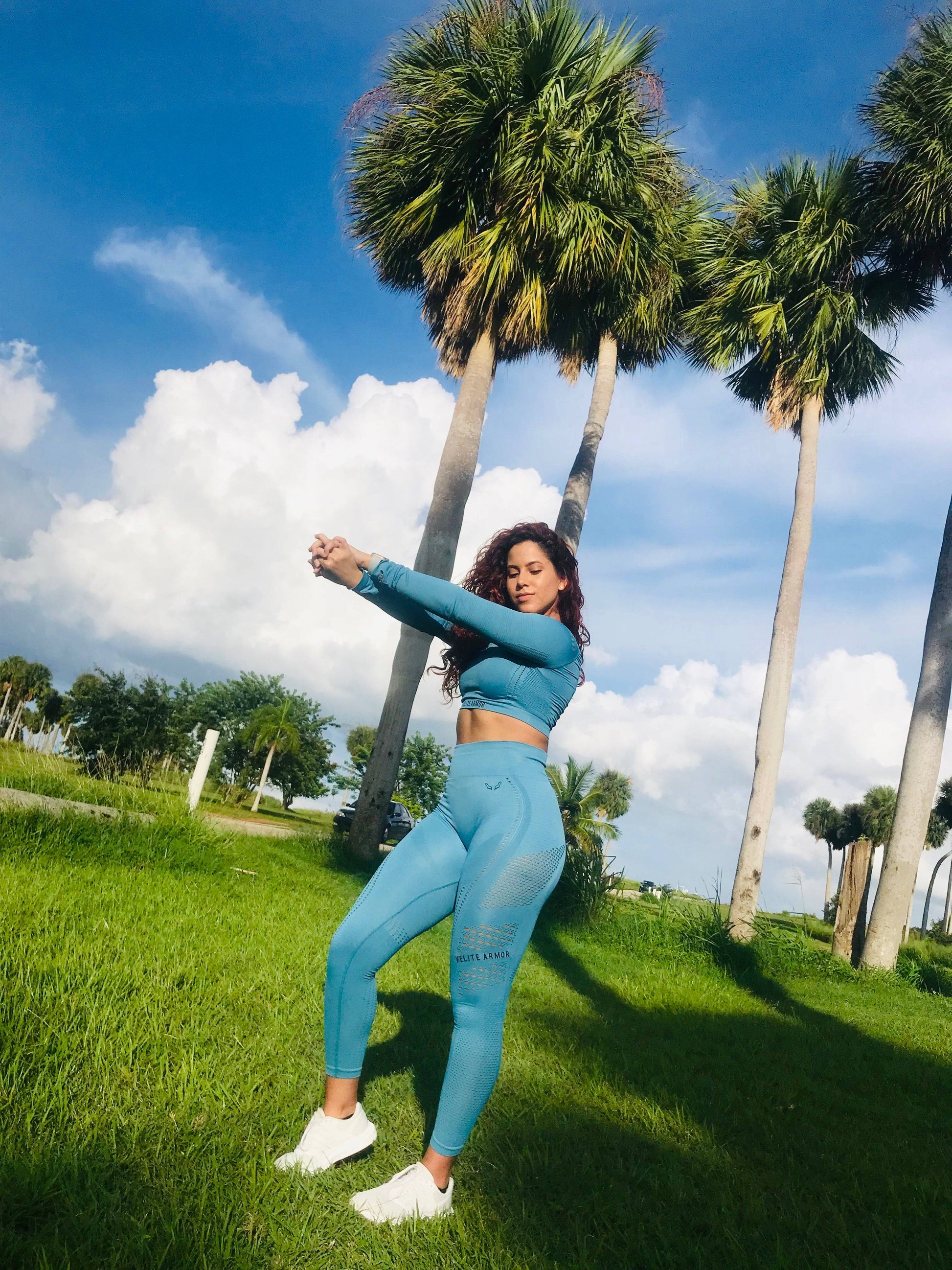 Woman stretching outdoors in a tropical setting, wearing a sky-blue seamless athletic set from Velite Armor. The outfit includes a long-sleeve crop top with breathable mesh details and high-waisted leggings designed for comfort and flexibility. Perfect for active lifestyles and outdoor workouts.