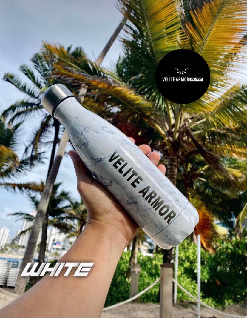 Velite Armor (ELITE) Water Bottle 17oz in white marble finish, thermal insulated, held in front of palm trees and a beach view.