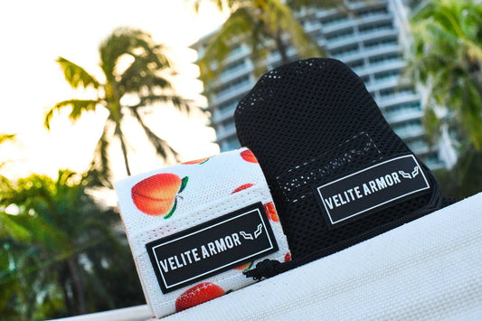 Velite Armor wrist wraps displayed in an outdoor setting with palm trees and a modern high-rise building in the background. One wrist wrap features a vibrant peach design on a white background, while the other is sleek black with a breathable mesh texture. These high-performance wrist wraps provide support and stability for weightlifting and intense training sessions.