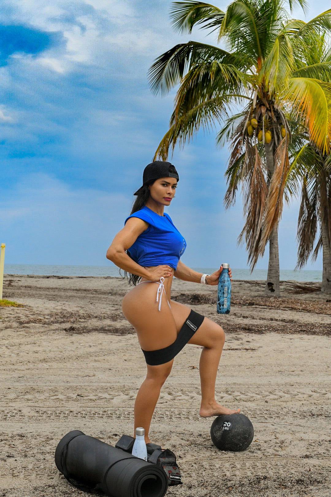 Mujer fitness usando una banda de resistencia de Velite Armor, posando en la playa con equipo de entrenamiento y palmeras de fondo.