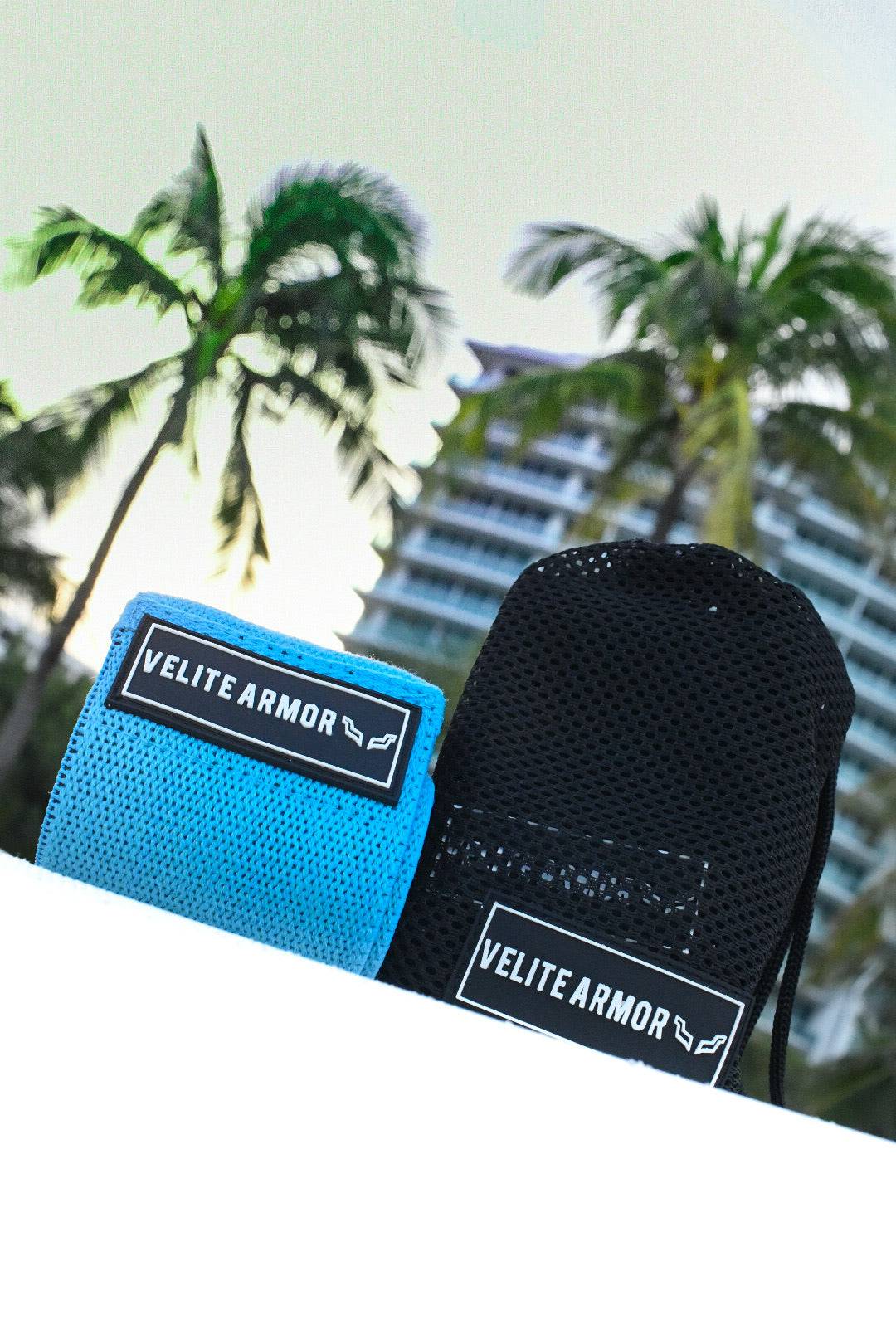 Velite Armor blue and black wrist wraps and lifting straps against a tropical backdrop with palm trees and a modern high-rise.