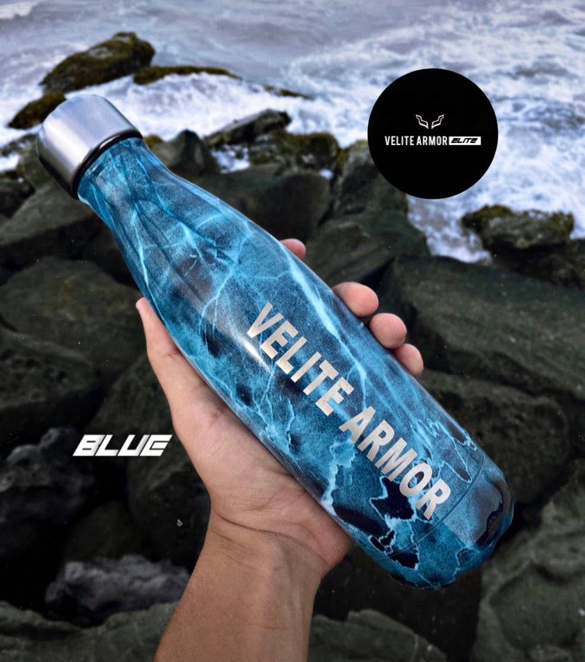 Velite Armor (ELITE) Water Bottle 17oz in blue marble pattern, stainless steel, displayed near ocean rocks for a rugged aesthetic.