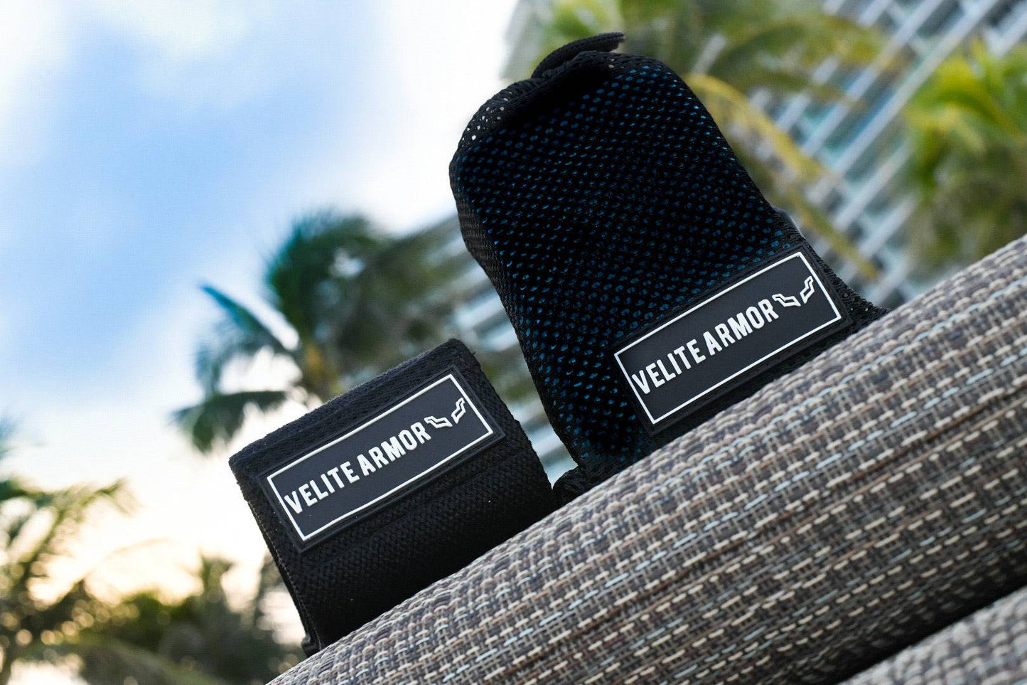 Velite Armor black wrist wraps and lifting straps on an outdoor surface with palm trees and a modern building in the background.