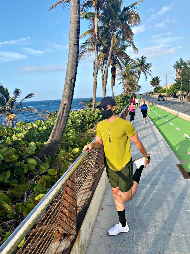 Athletic man stretching his quadriceps while holding onto a railing on a scenic coastal path lined with palm trees. He is wearing a yellow Velite Armor performance t-shirt, black shorts, white sneakers, and a face mask. Other runners and walkers are seen in the background, enjoying the ocean breeze. A perfect setting for outdoor training and an active lifestyle.