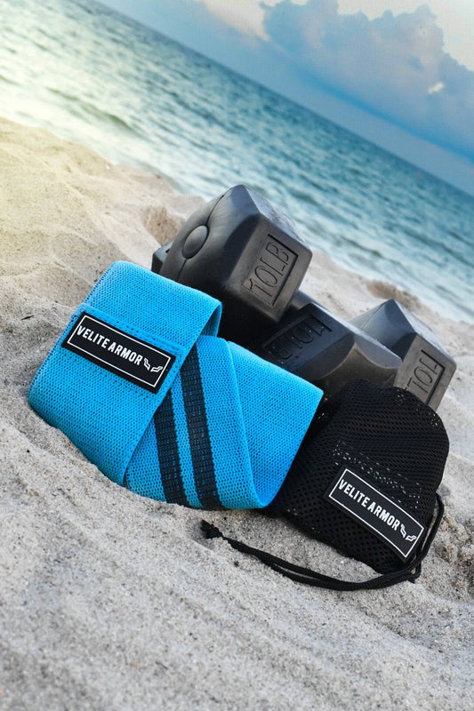 Velite Armor resistance band and wrist wraps on the sand next to dumbbells, with the ocean in the background.