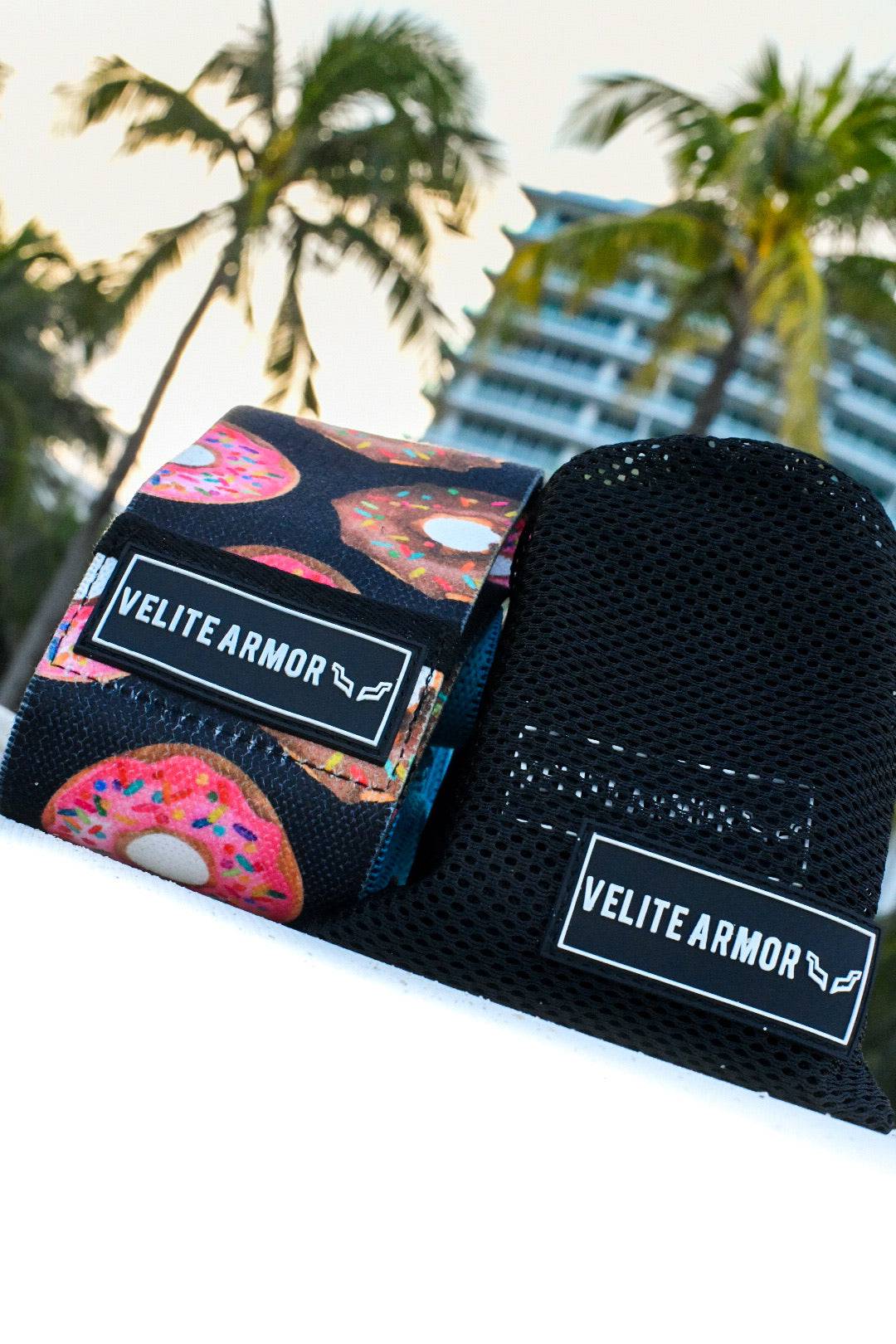 Velite Armor wrist wraps with a playful donut pattern, displayed alongside a black mesh carrying pouch against a backdrop of palm trees and a modern high-rise building. Stylish and functional accessories for weightlifting and strength training.